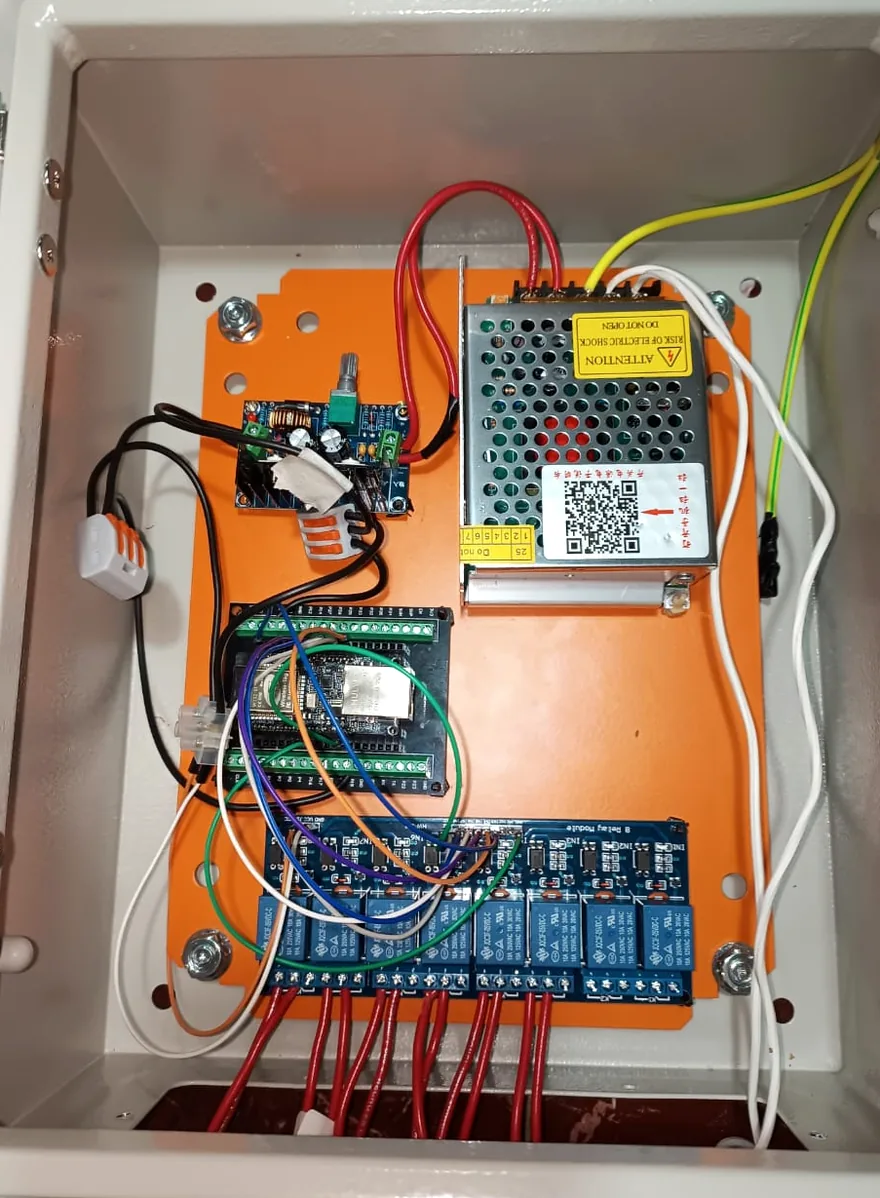 Caja de control terminada dentro de su gabinete: placa de control, fuente de poder y bloque de ocho relés, cableados sobre una placa de soporte naranja.